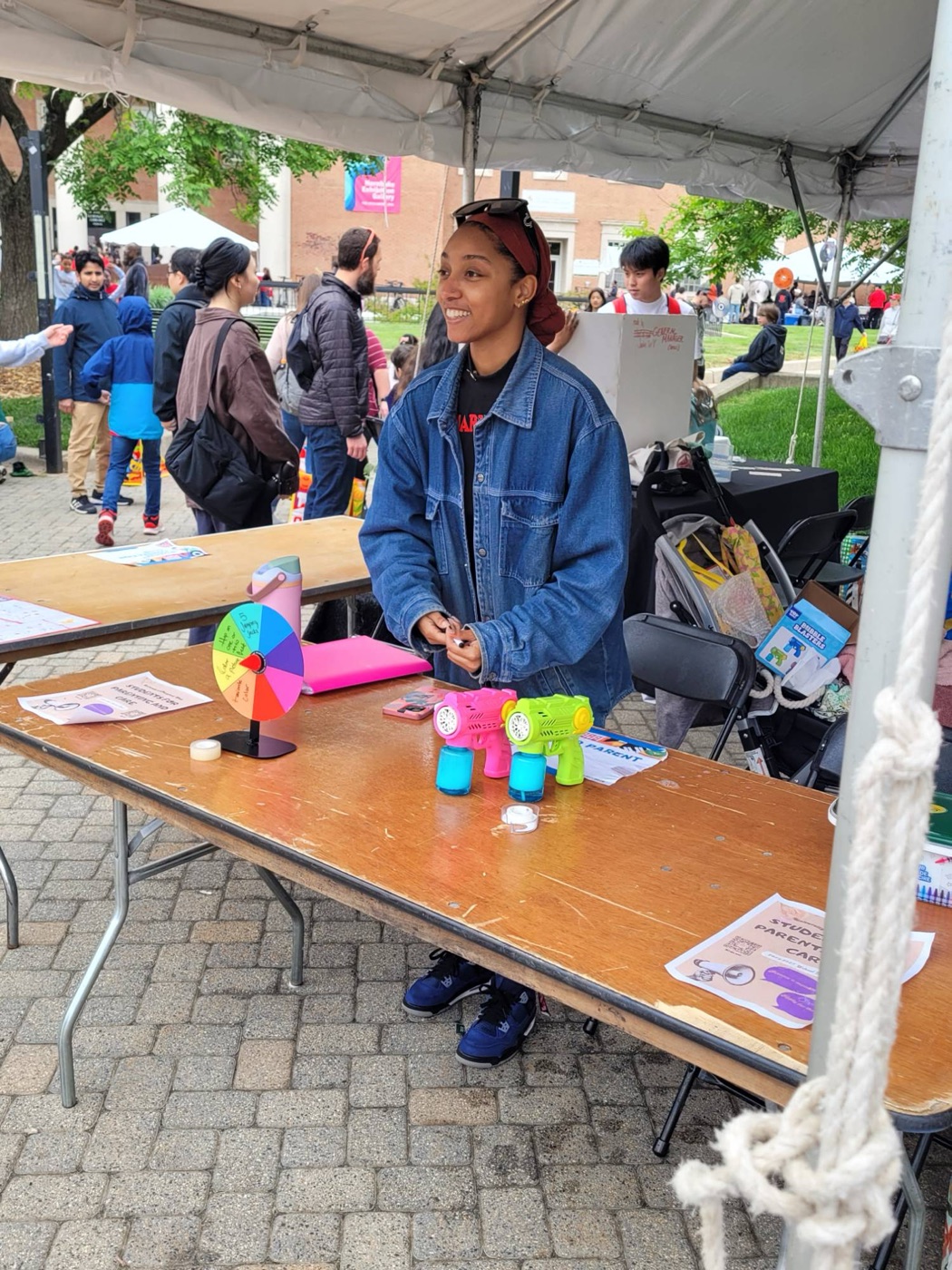 Amaya smiling behind the Students for Parenting and Care table at Maryland Day, with a prize wheel and bubble blasters for kids